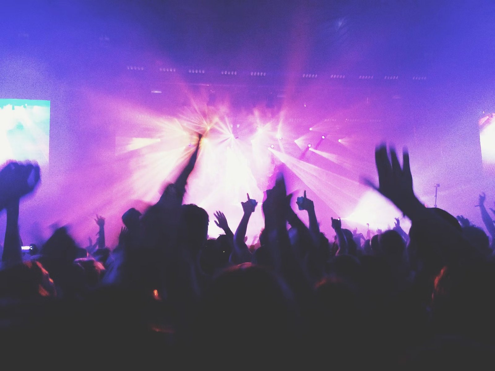 A crowd lifts hands beneath purple beams and stage haze.